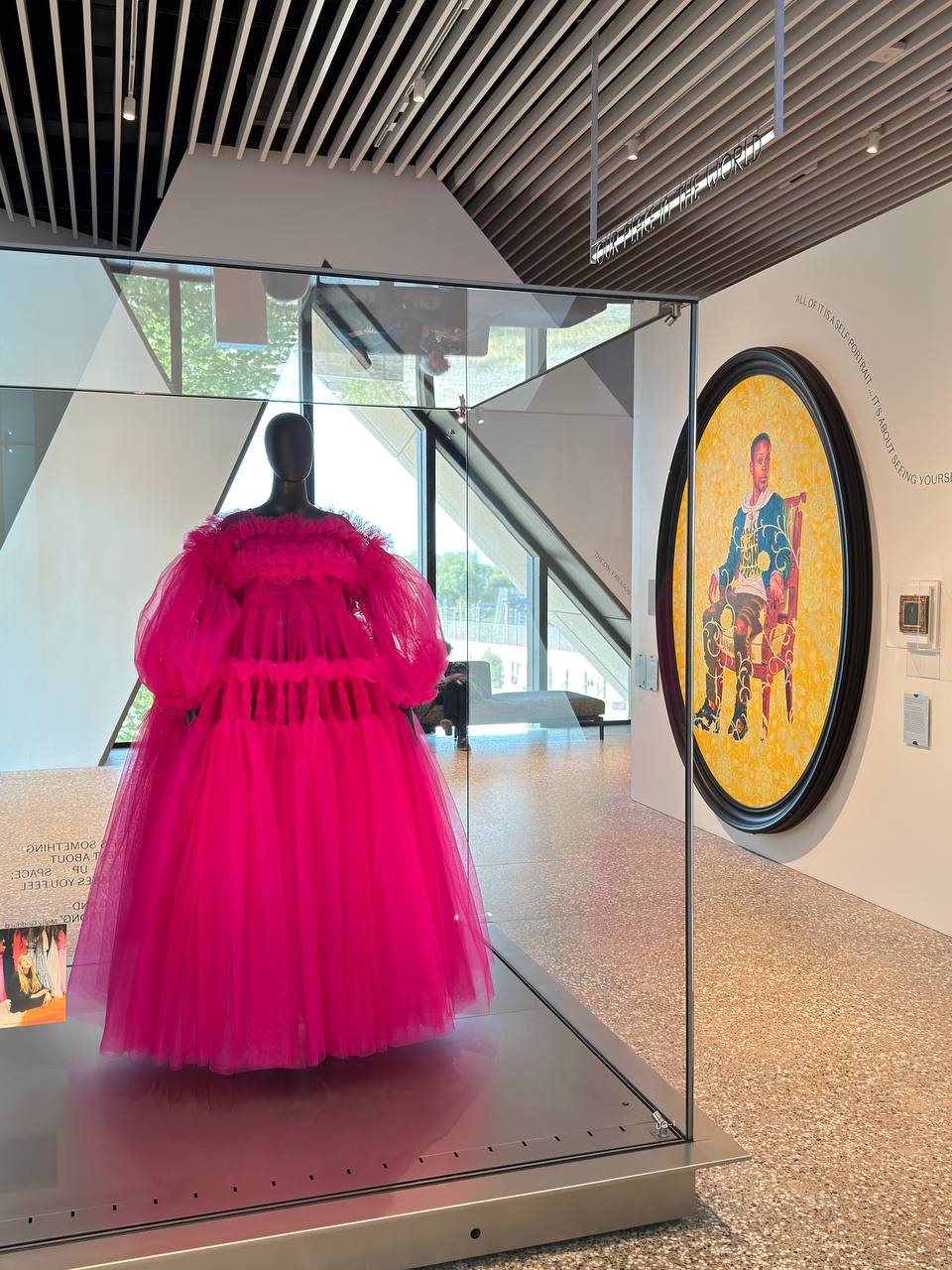 A pink tulle Molly Goddard dress in a glass case beside a large round portrait of a young man seated on a carved chair.