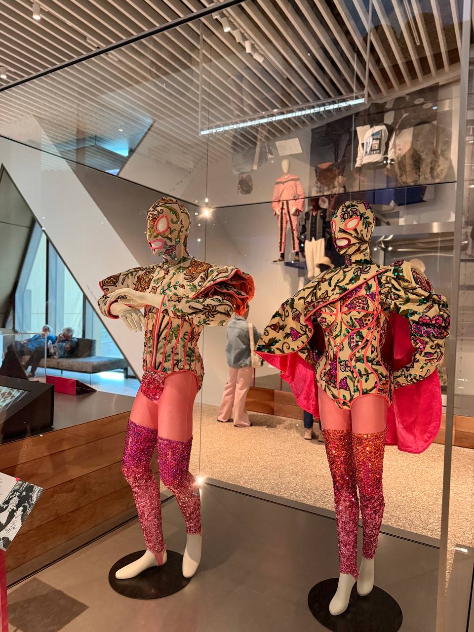 Two sequinned stage costumes with embroidered full-face masks and pink capes stand in a glass case.