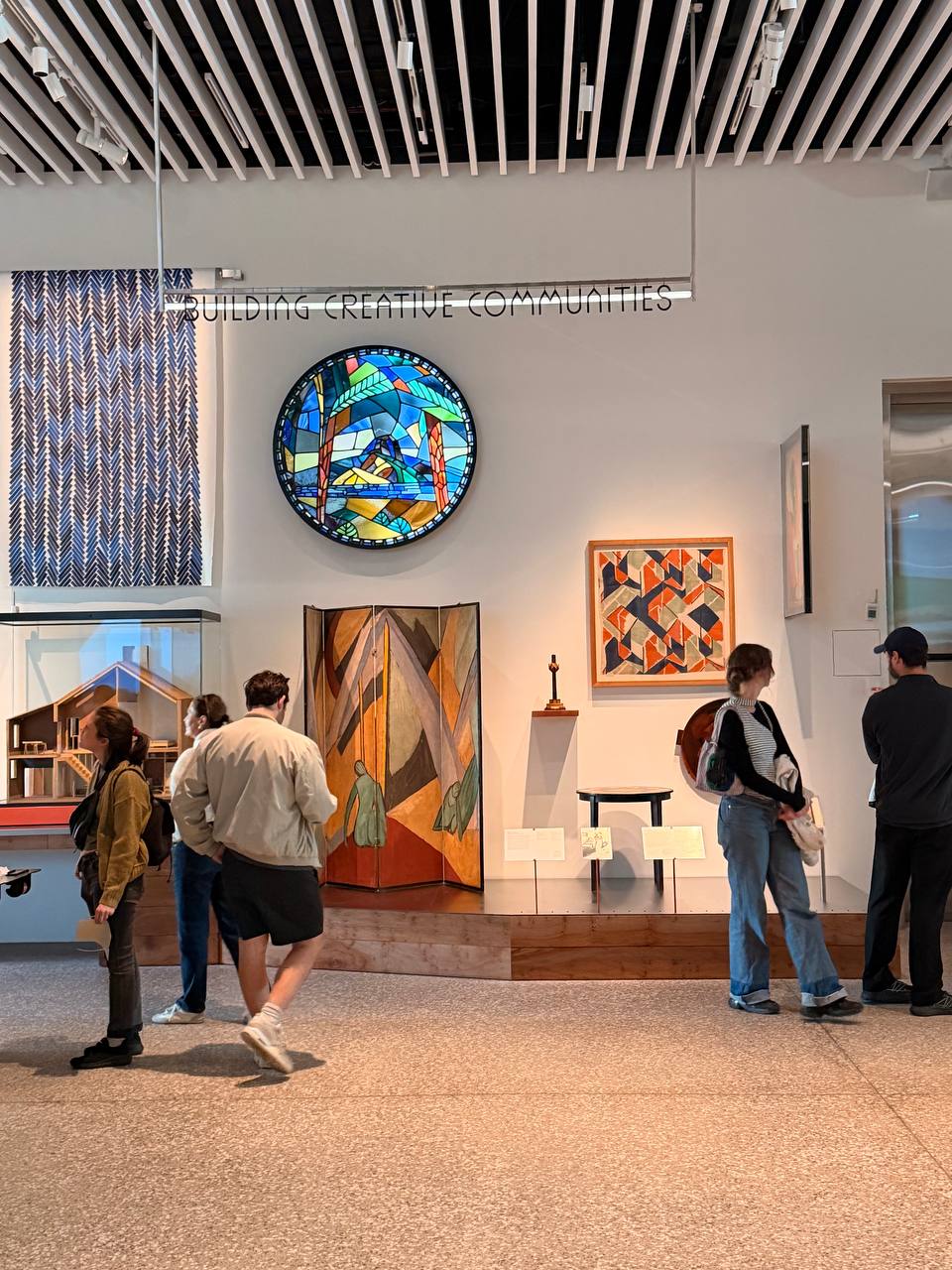 Visitors walk past a wall headed Building Creative Communities, with a stained-glass roundel above a painted folding screen and a framed textile.