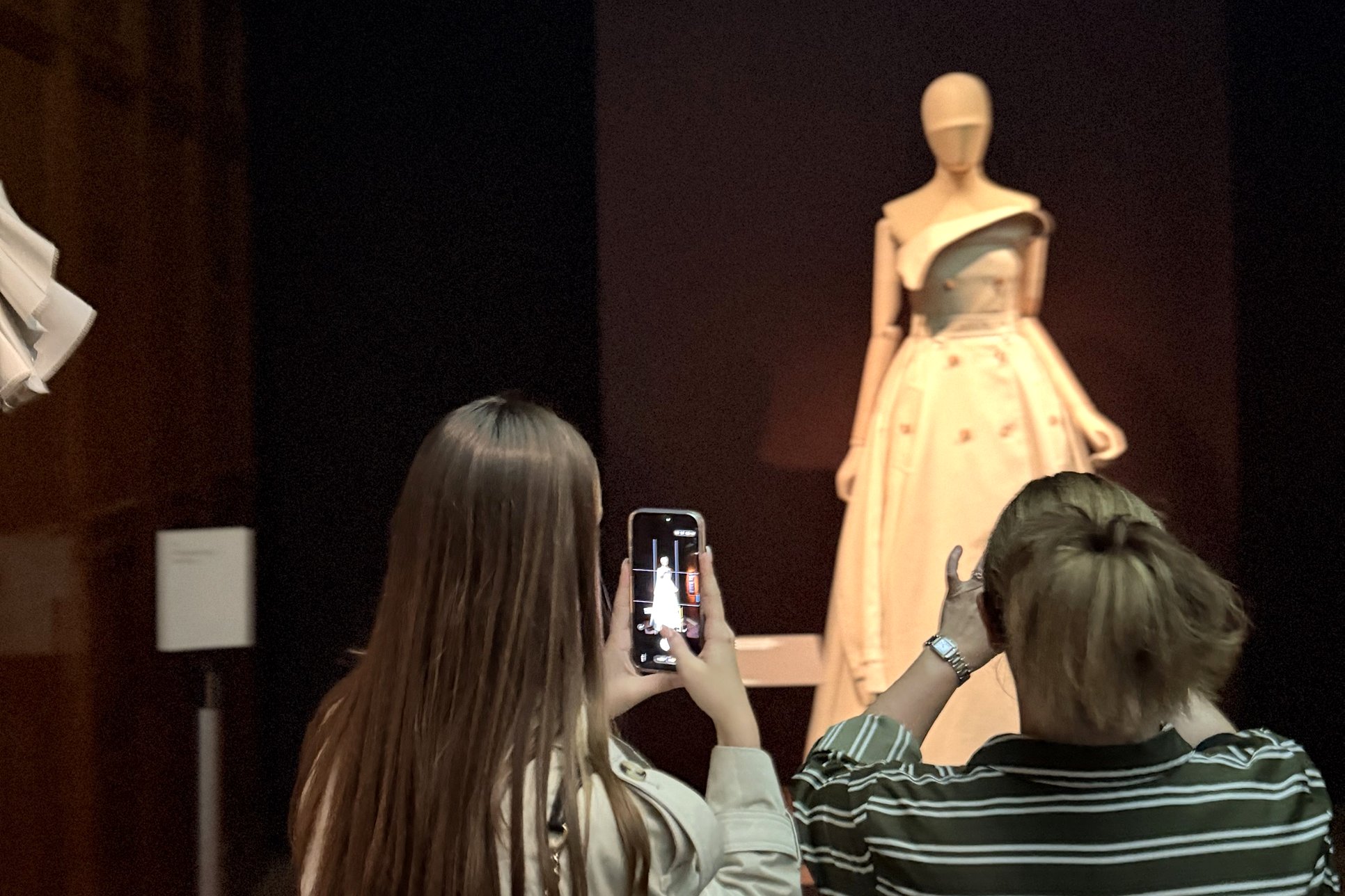 Visitor in a beige trench photographing Riccardo Tisci's strapless trench ballgown at the end of the Burberry display at the V&A.