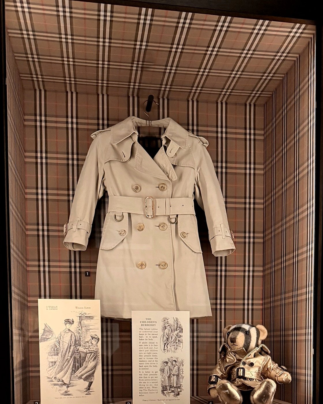 Small beige child’s trench coat hanging in a case lined with Burberry check, above two printed advertisements and a teddy bear in a gold trench.