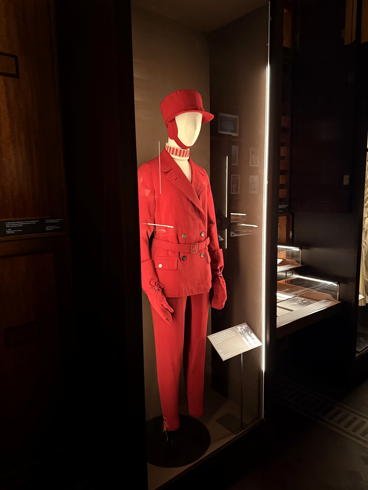 Red Burberry ski suit with matching cap, belted jacket and gloves on a mannequin in a lit glass case.