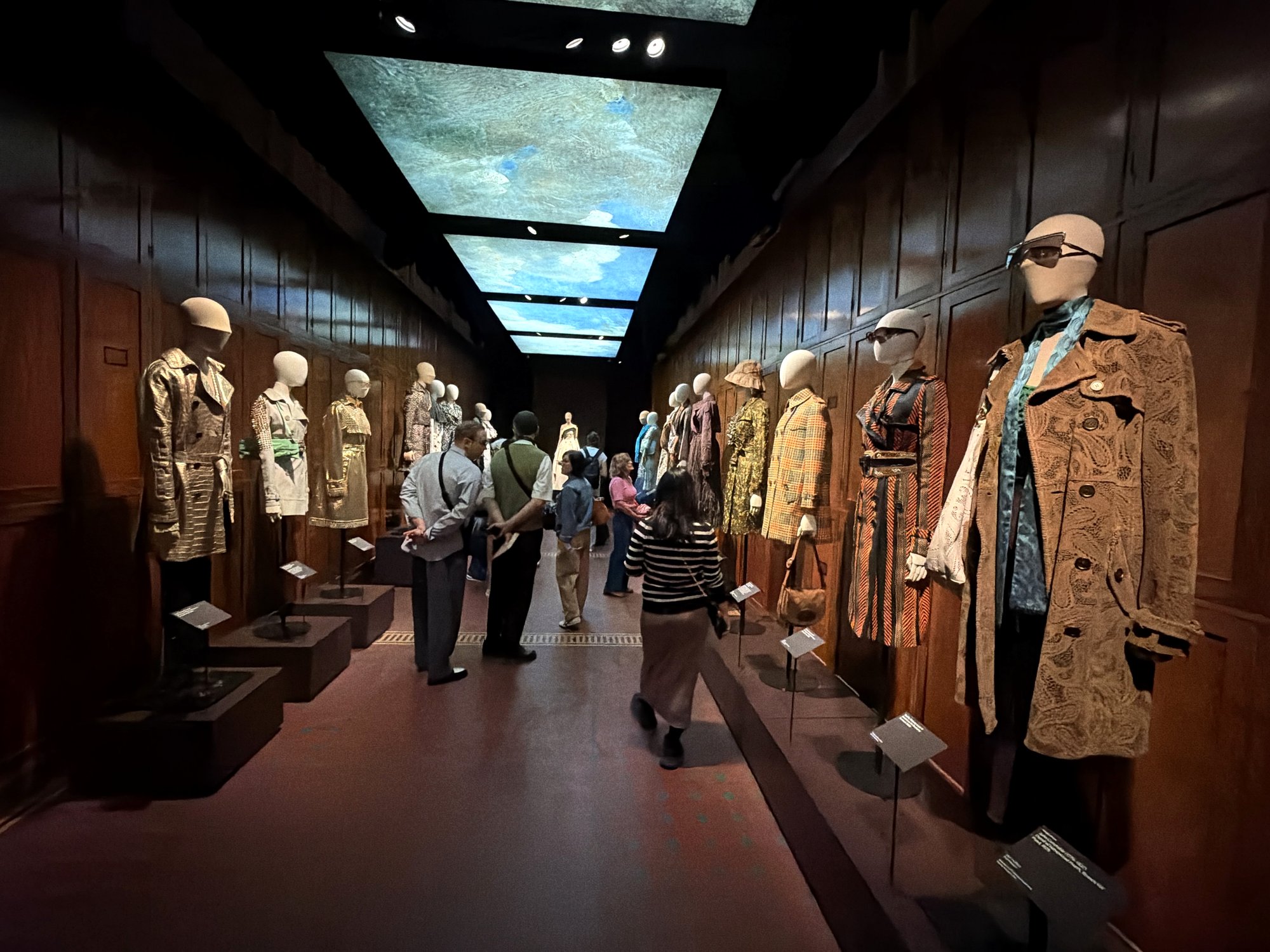 Long wood-panelled corridor with runway trench looks on plinths along both walls, visitors walking between them and glowing sky panels overhead.