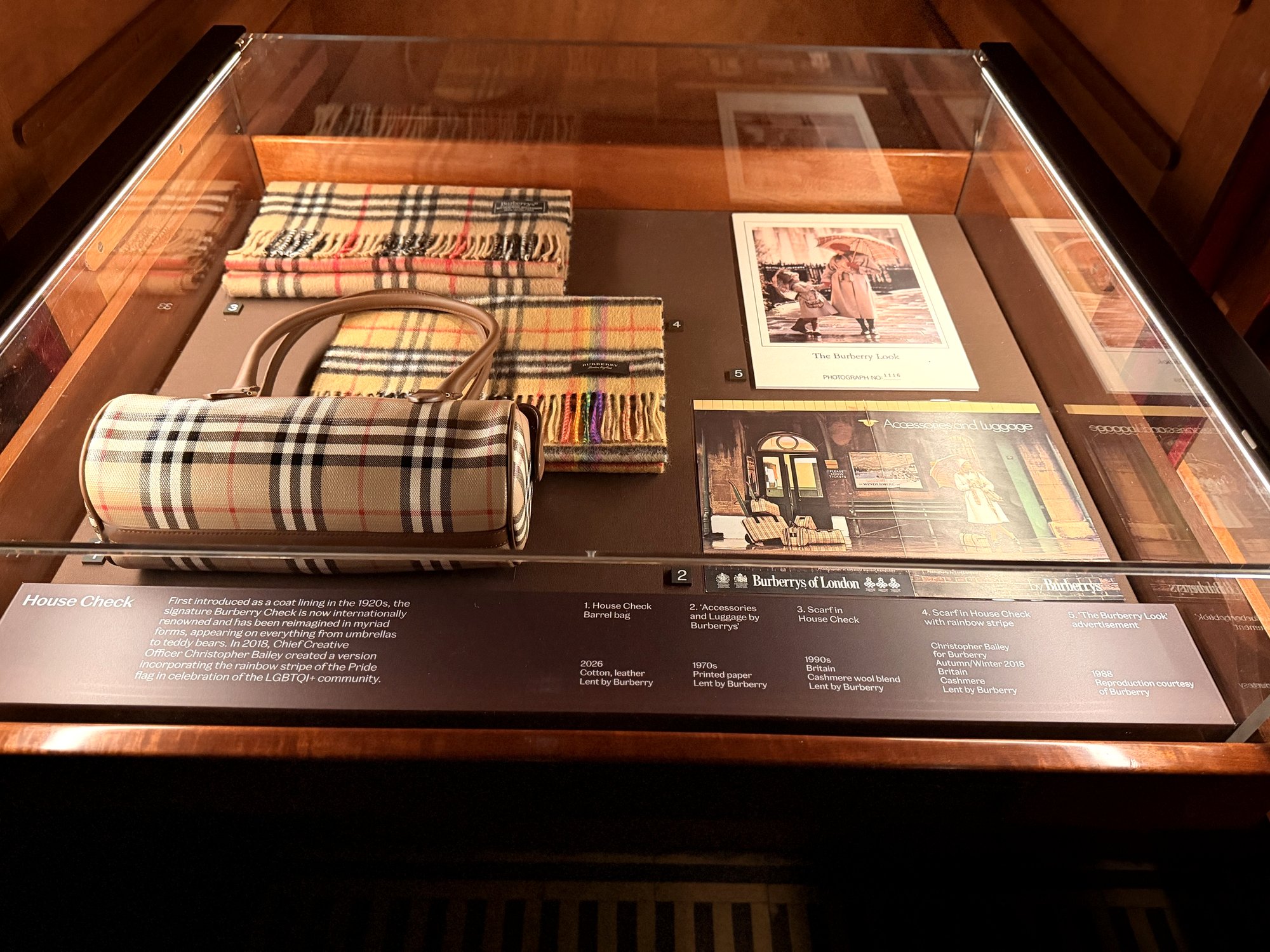Glass drawer case holding a Burberry check barrel bag, two check scarves and two printed advertisements, with a label panel below.