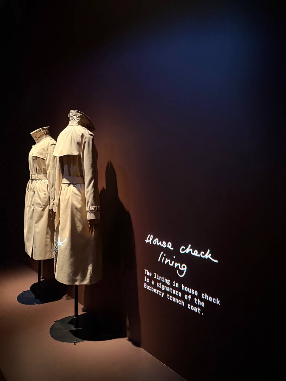 Two beige trench coats on stands in a dark room, beside handwritten projected text reading House check lining.