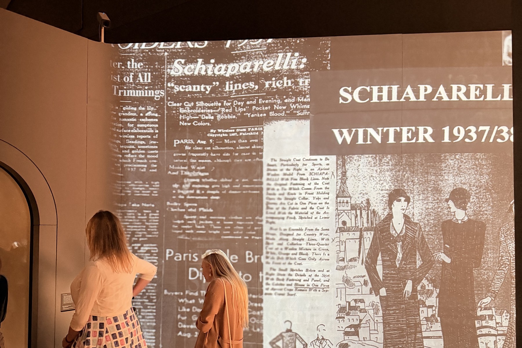 Two visitors stand with their backs to us before a large projection reading ‘Schiaparelli — Winter 1937/38’ at the V&A.