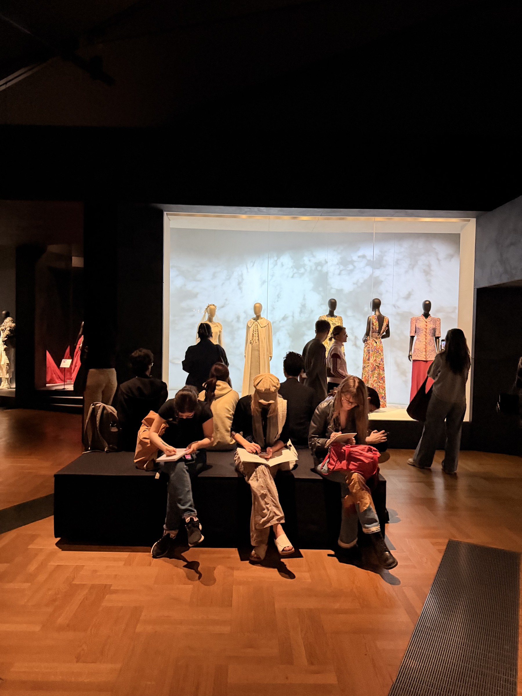 Young visitors sit sketching in front of a lit case of Schiaparelli mannequins at the V&A.