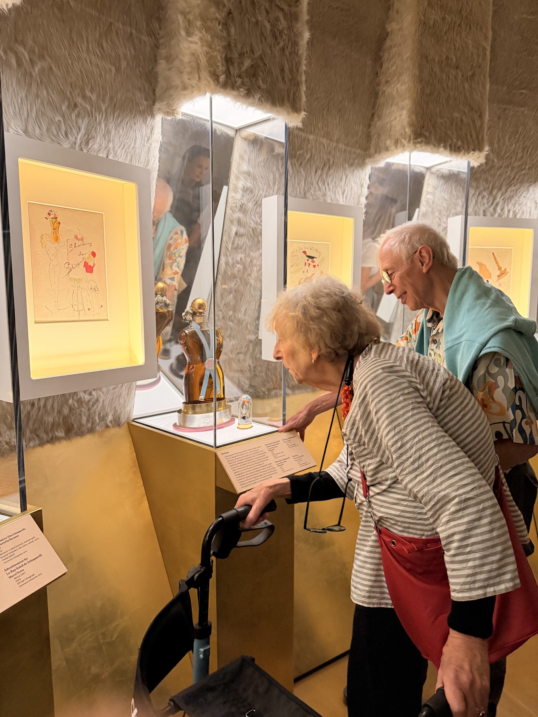 An older couple lean in to read the label beside the ‘Shocking’ perfume bottle in a mirrored, fur-lined case at the V&A.