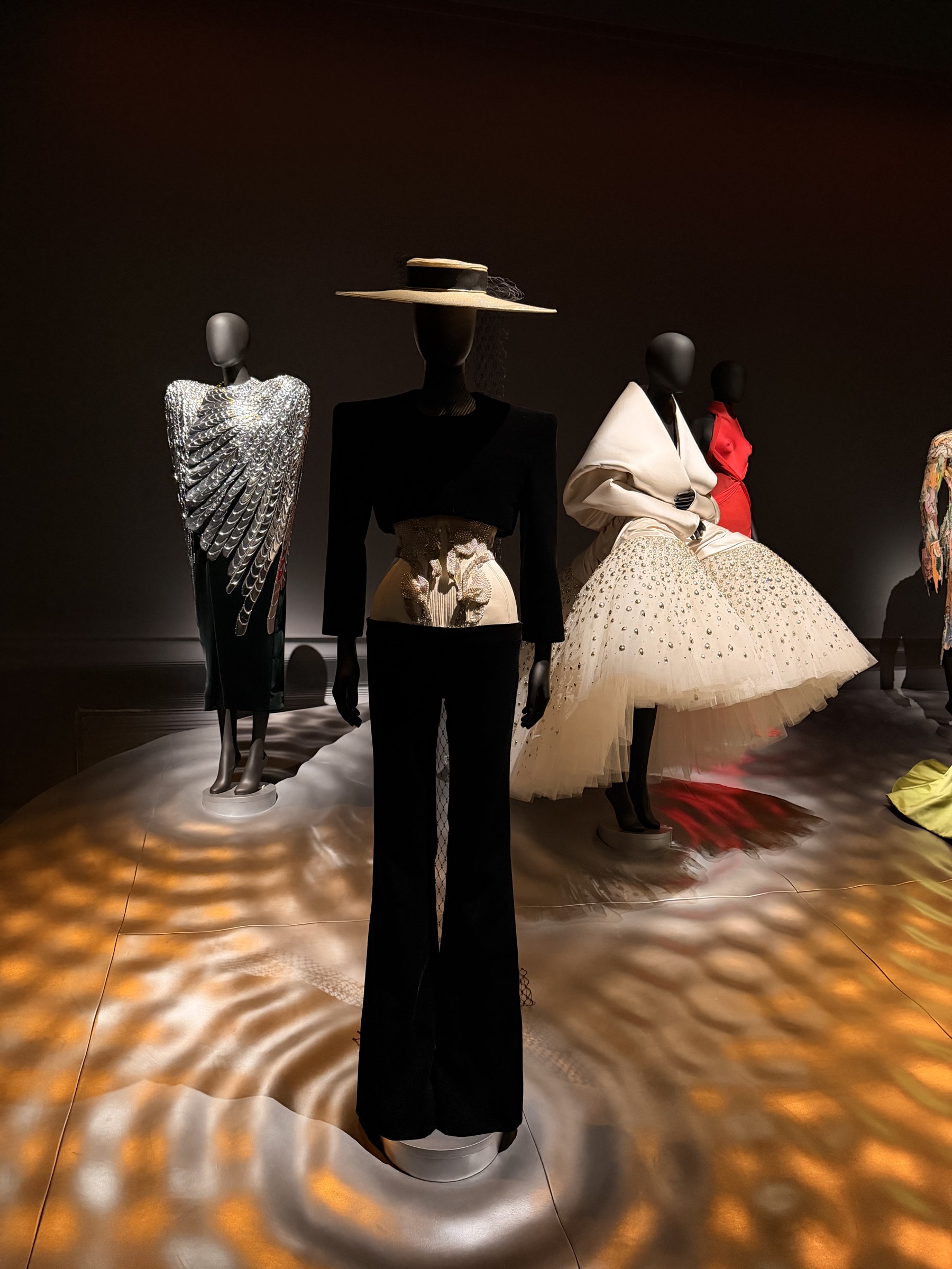 A black Schiaparelli jacket, straw hat and sculpted bodice at the centre of a lit room, with a tulle gown and a winged dress alongside.