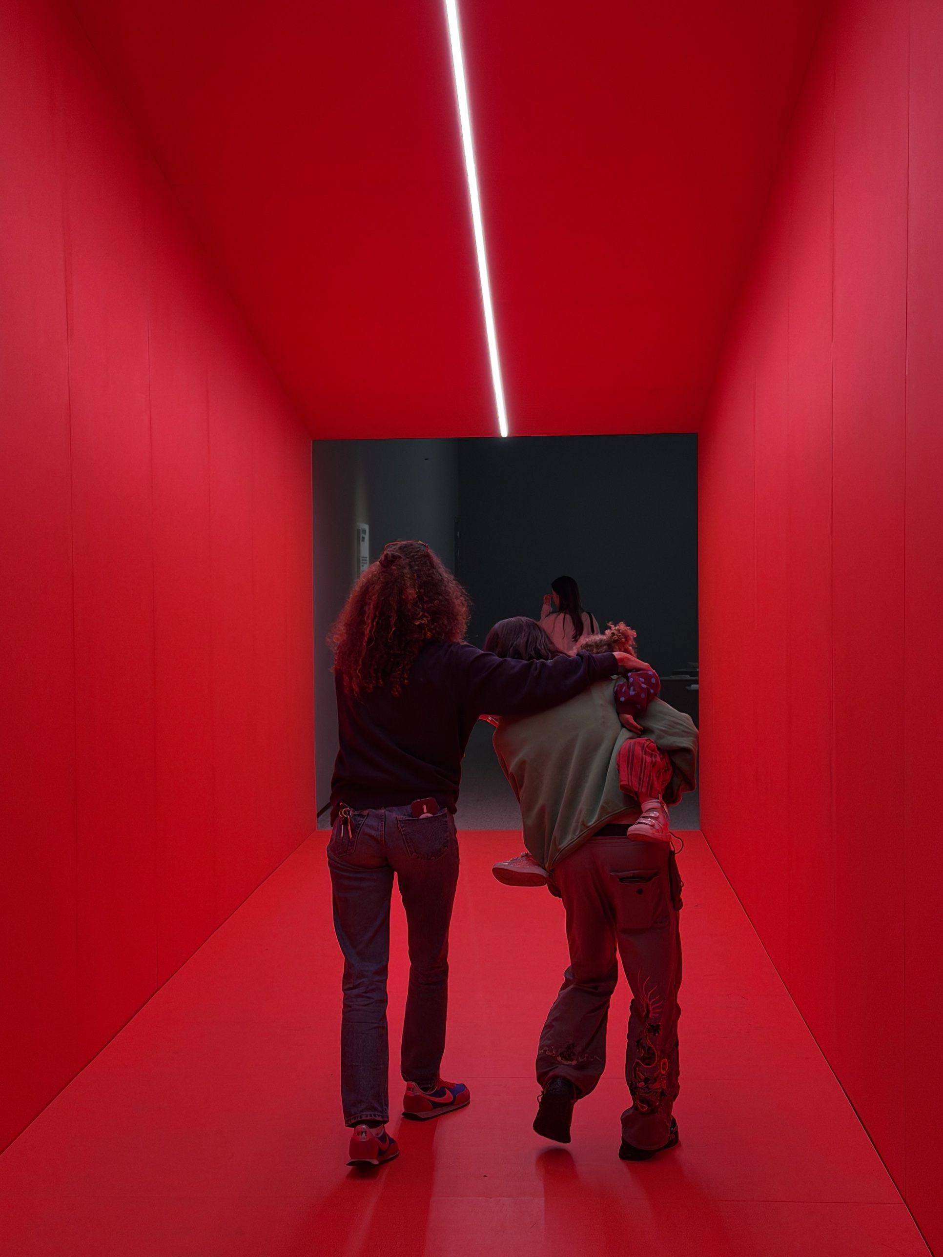 Two adults walk away down a glowing red corridor at the Design Museum, one carrying a small child toward the dark gallery beyond.