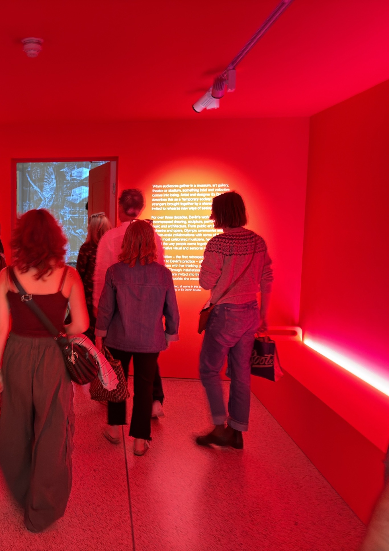 Visitors seen from behind in a red-lit room, facing a wall text that describes the audience as a temporary society.
