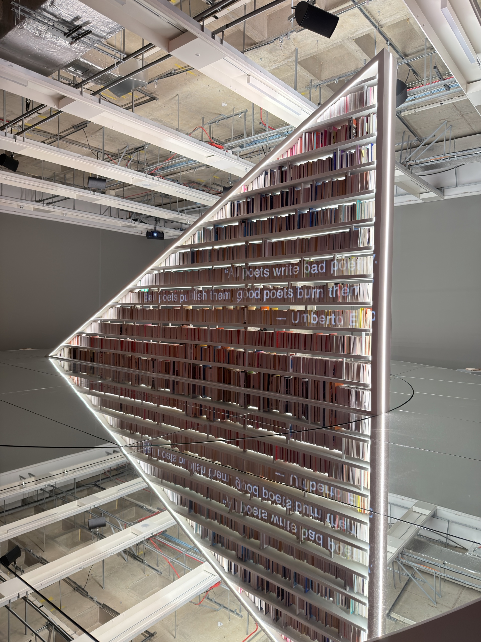 A towering triangular wall of colour-sorted books reflected in a mirrored floor, crossed by a line of illuminated text.