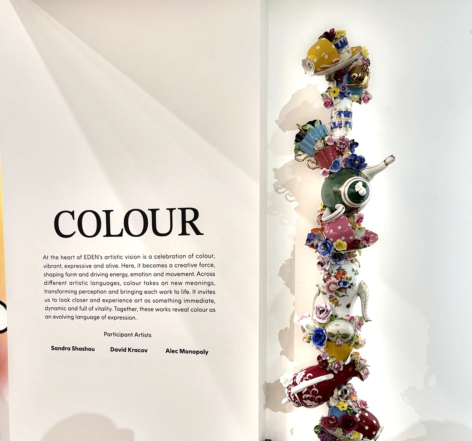 A gallery wall headed COLOUR with a curatorial paragraph and three artist names, beside a column of stacked porcelain teapots, cups and flowers