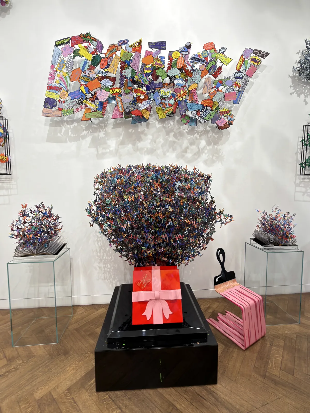 Cut-metal butterflies rising out of a lacquered red gift box on a black plinth, flanked by two smaller butterfly works on glass plinths