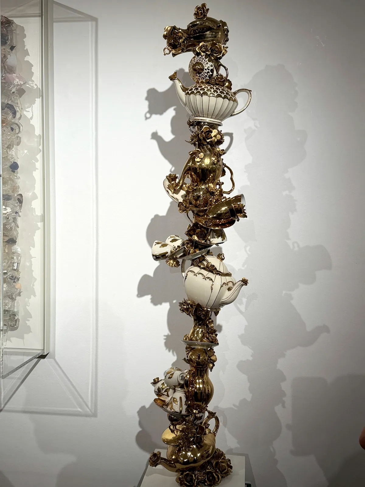 A tall column of gilded teapots, cups and porcelain roses stacked into a totem, casting a second column in shadow on the wall