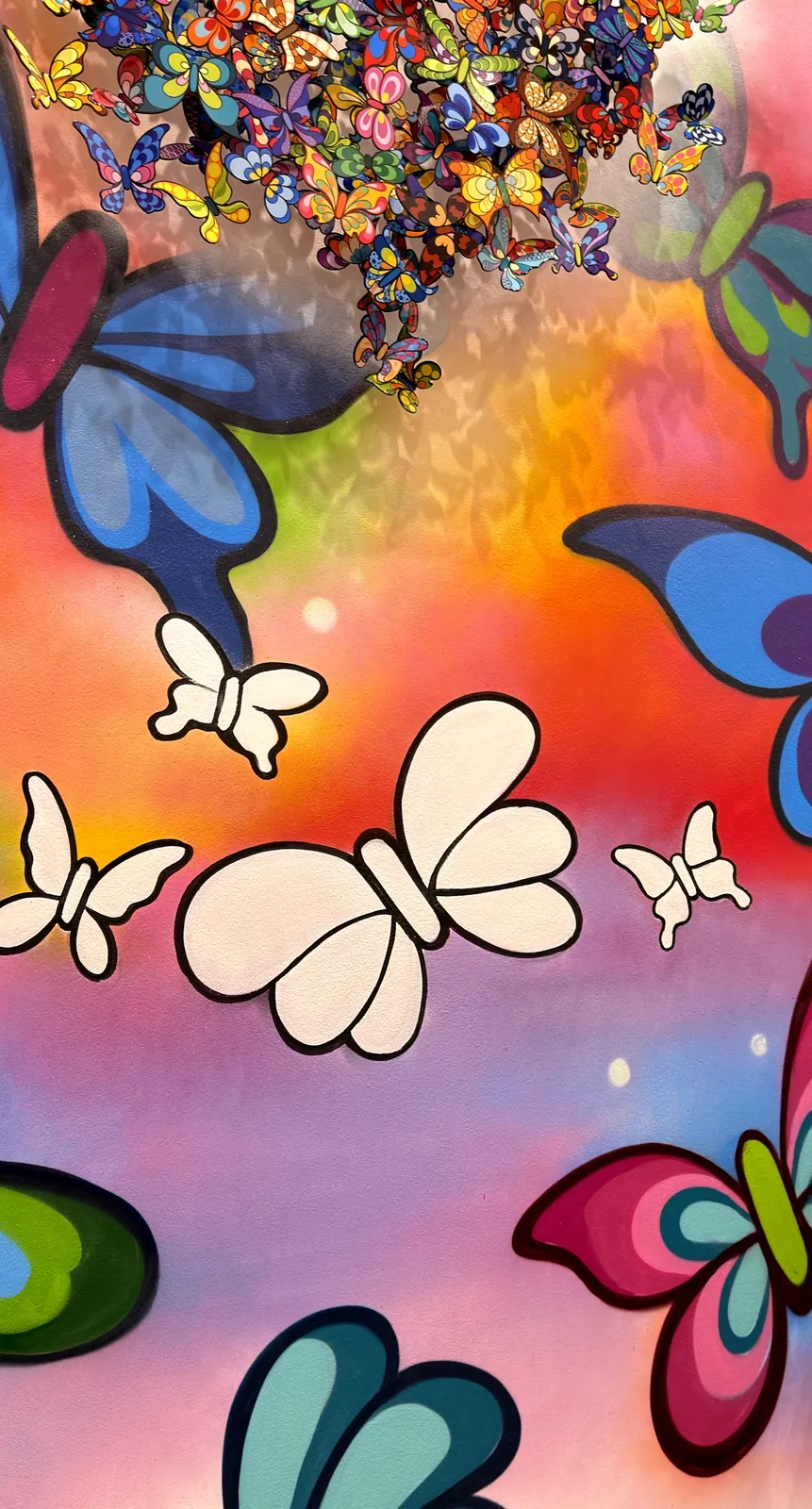 A wall of butterflies drawn in outline over a sprayed pink, orange and violet ground, some filled in with colour by visitors