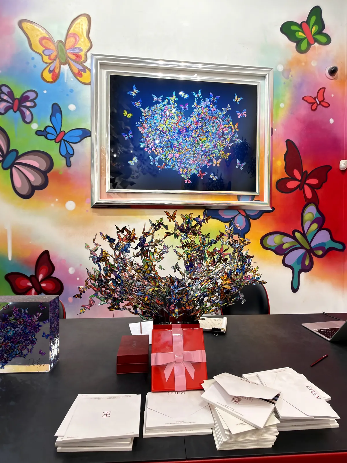 A gallery desk stacked with headed envelopes and a ribboned red box, in front of a wall painted with the gallery's butterfly motif