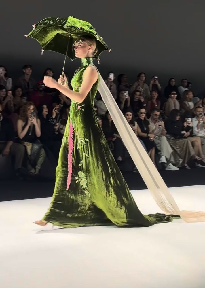 A model in a green velvet gown holding a matching embroidered parasol with a long cream chiffon train