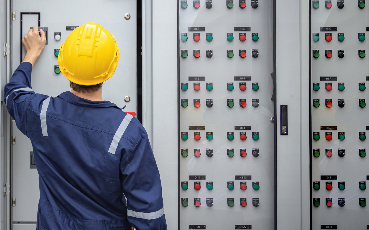 Industrial worker in a yellow helmet operating control panel with buttons and switches.