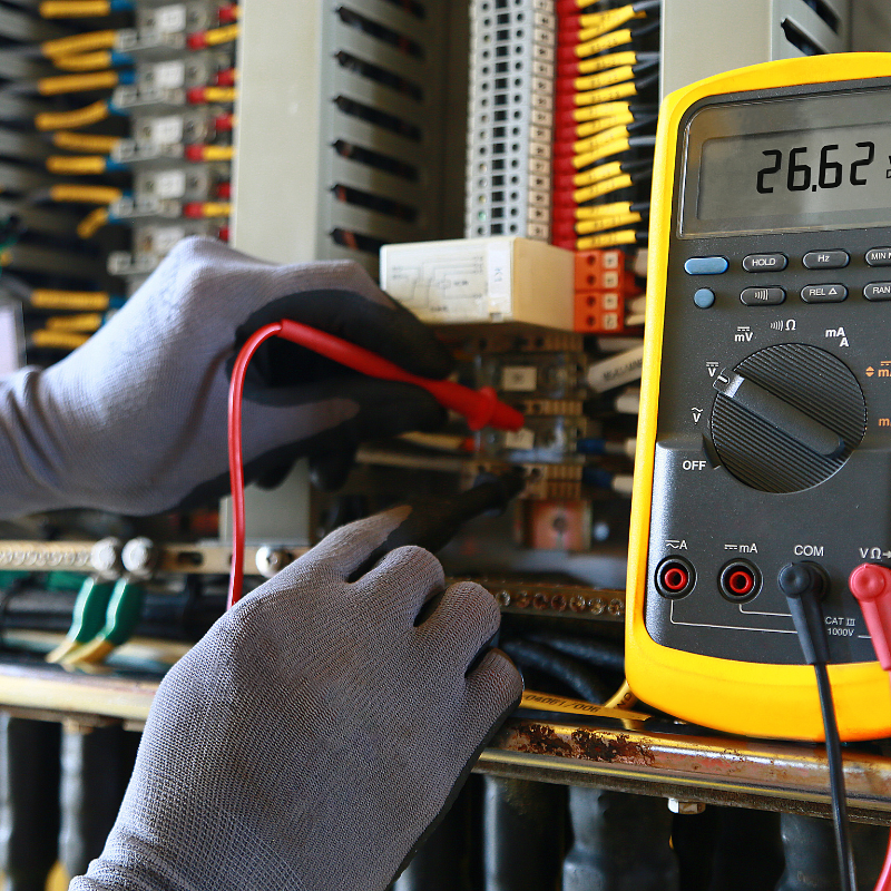 Electrician wearing gloves using a digital multimeter to test voltage in an electrical panel.