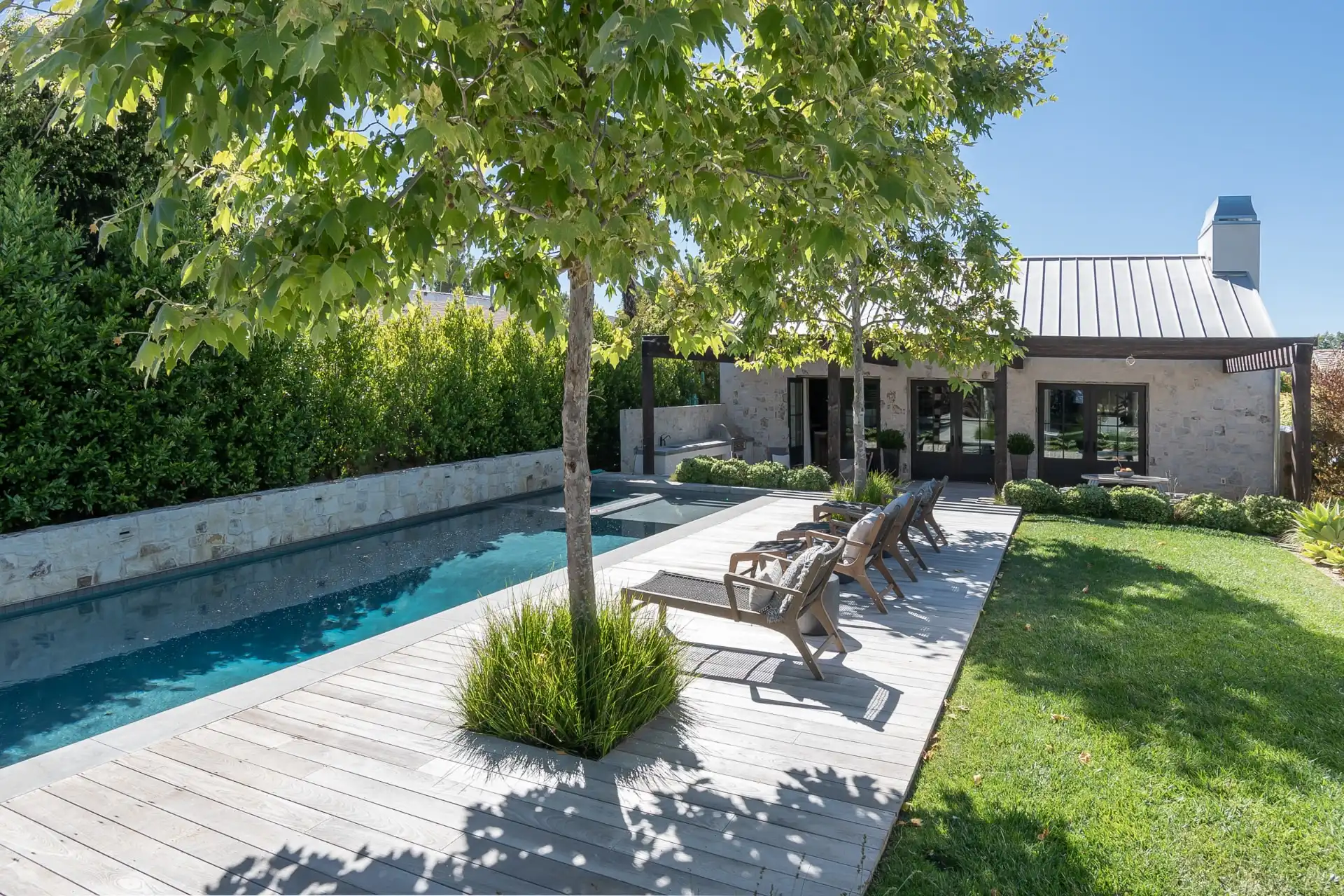 Backyard swimming pool beside a landscaped lawn, patio seating and a stone-clad house.
