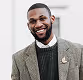 Smiling man in a gray checkered blazer and black sweater standing outdoors.