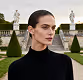 Woman with dark hair wearing a black turtleneck sweater standing outdoors with trimmed bushes and statues in the background.