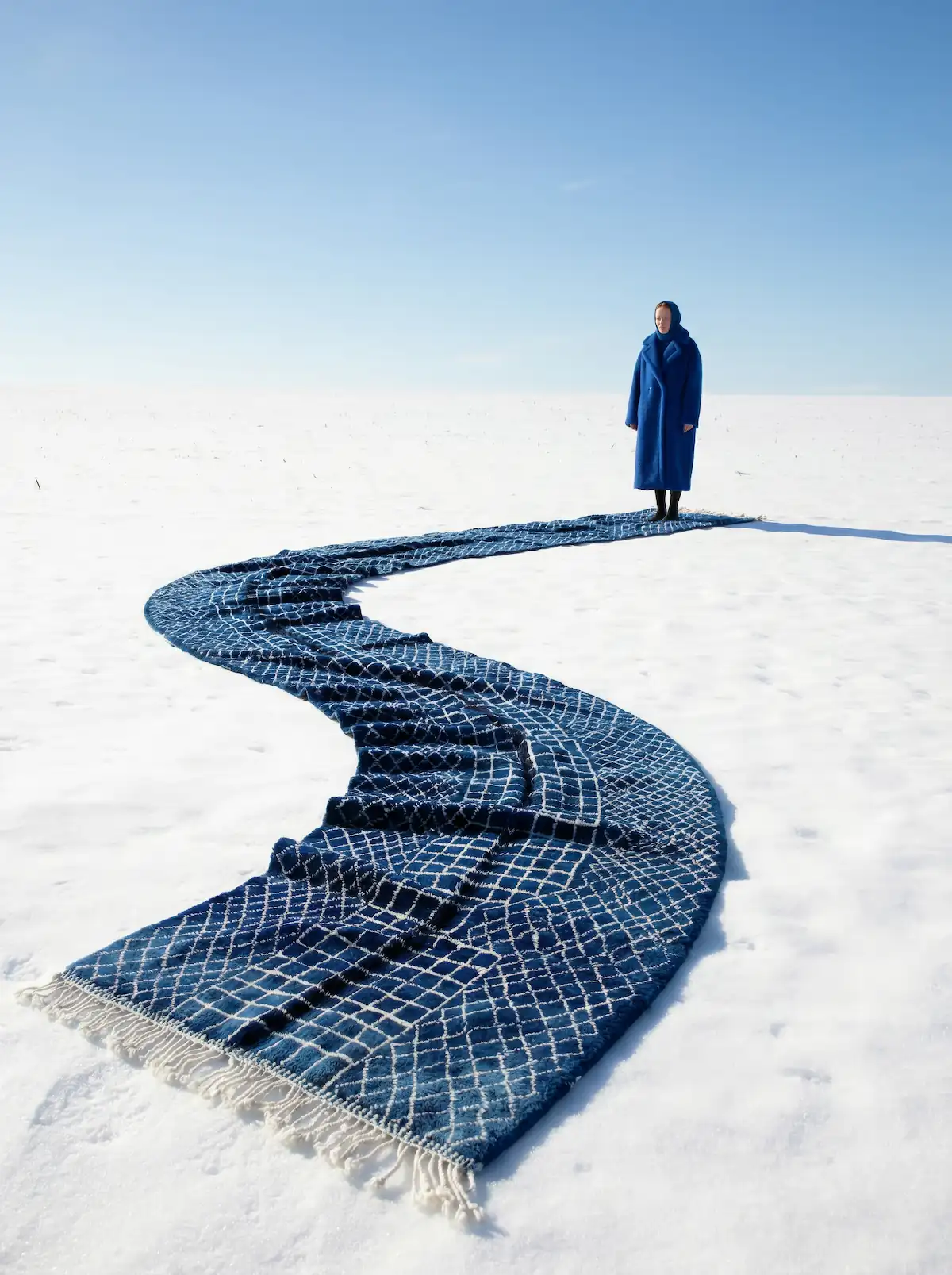 Woman in a blue coat standing at the end of a winding blue Moroccan rug on snow