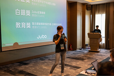 A woman speaking with a microphone in a conference room with a presentation projected on the wall.