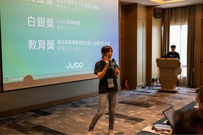 A woman speaking with a microphone in a conference room with a presentation projected on the wall.