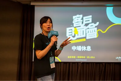 女性講者在演講中，背景投影顯示中文文字與會議主題。