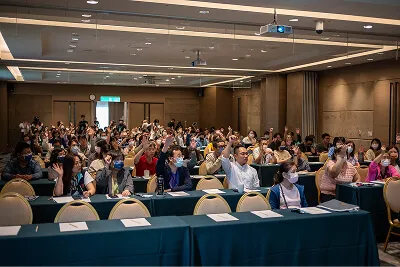 會議室內眾多戴口罩的與會者舉手互動，桌上擺有紙張和筆