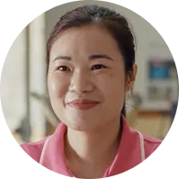 微笑的亞洲女性，穿著粉紅色衣服，背景為室內模糊場景。