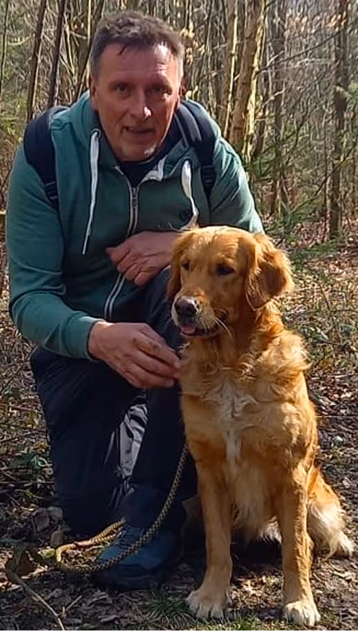 Man in a green hoodie kneeling beside a sitting golden retriever in a forest.