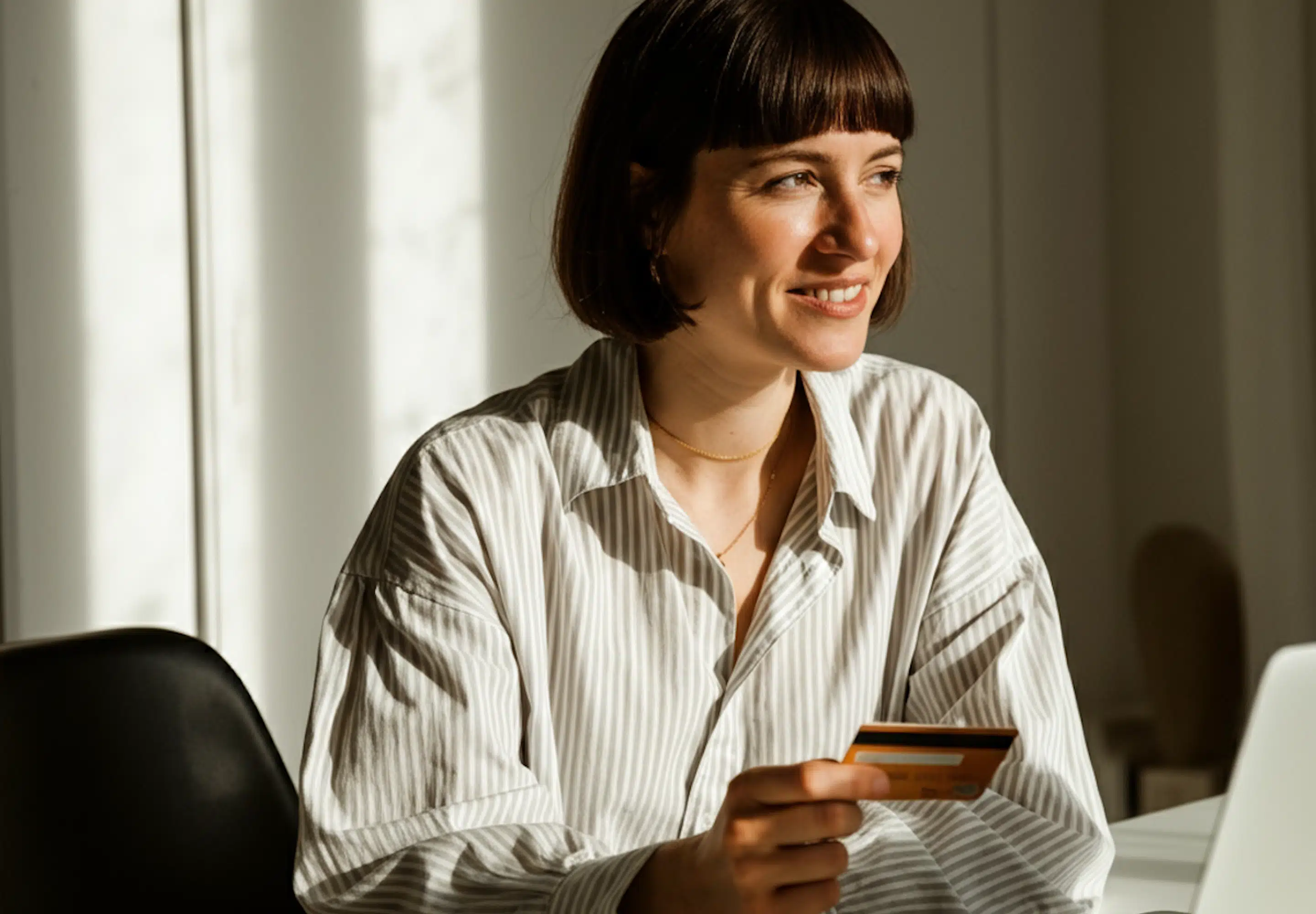 Smiling woman holding a payment card while working at a laptop in sunlight