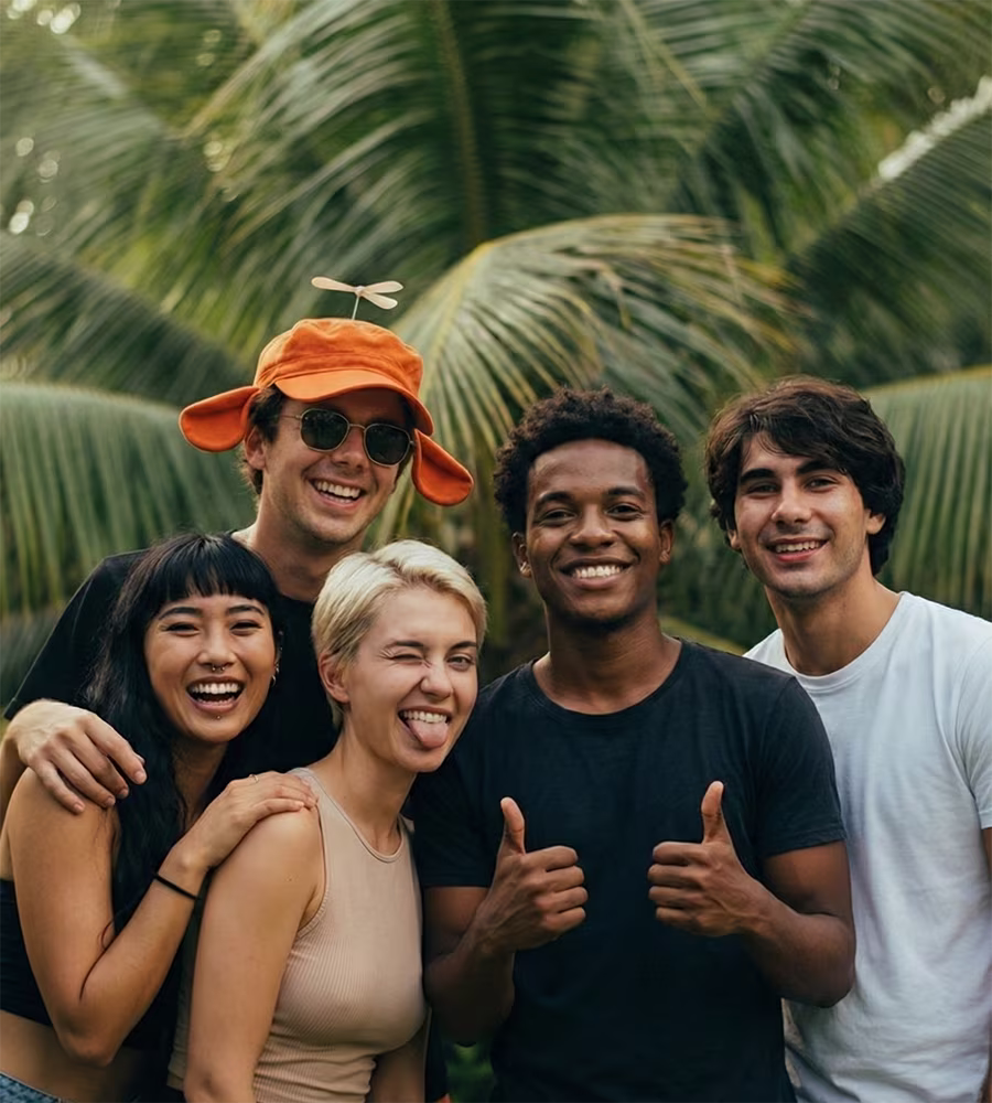 A cheerful group portrait of five young people posing playfully with thumbs-up gestures against a tropical background of palm trees.