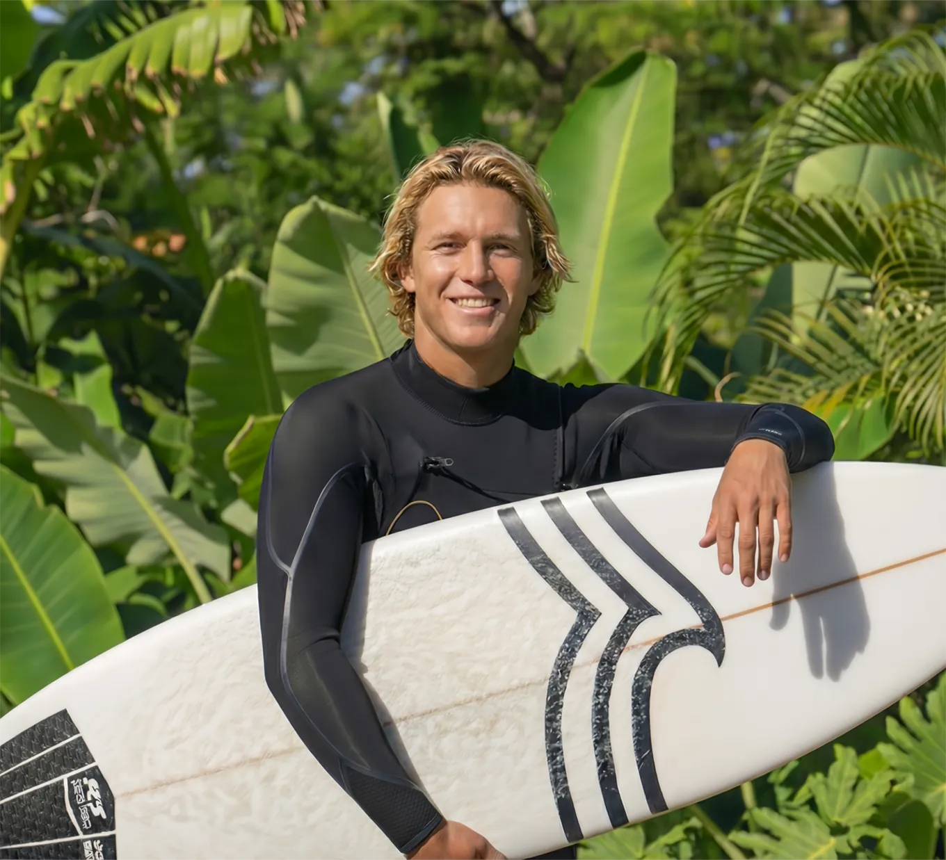 A portrait of a friendly man in a wetsuit posing with his surfboard in front of vibrant green palm leaves.