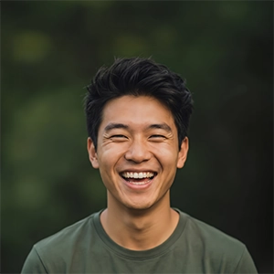 A man with dark, styled hair laughing and showing a bright smile, wearing a dark green t-shirt against a blurred forest background.