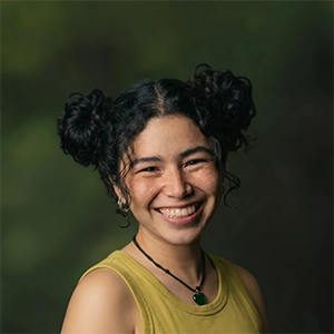 A smiling woman with freckles and dark hair styled in two buns, wearing a yellow tank top and a heart necklace against a blurred green background.