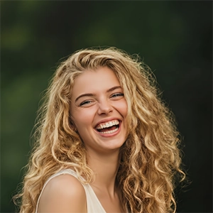 A joyful woman with long, curly blonde hair laughing heartily, set against a soft-focus natural green background.