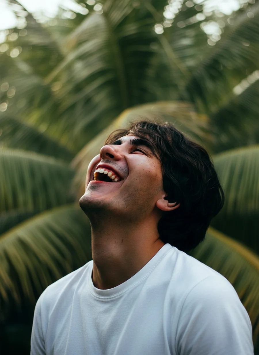 A man with dark hair laughing heartily with his head tilted back, set against a blurred green foliage background.