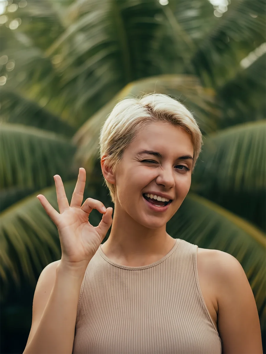 A woman with short blonde hair winking and making an "OK" hand gesture while smiling in a lush green outdoor environment.
