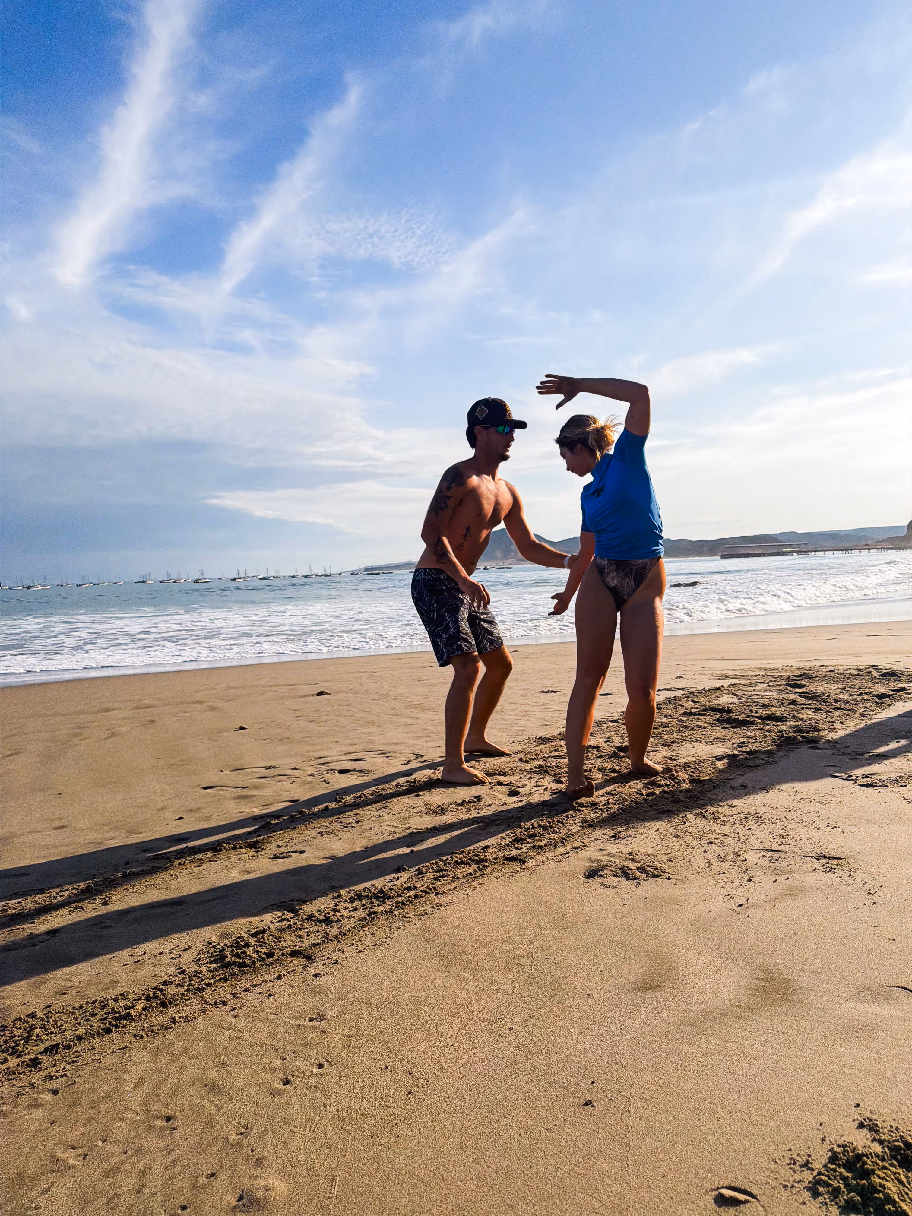 A man and woman on a beach with the ocean and boats in the background during a sunny day.