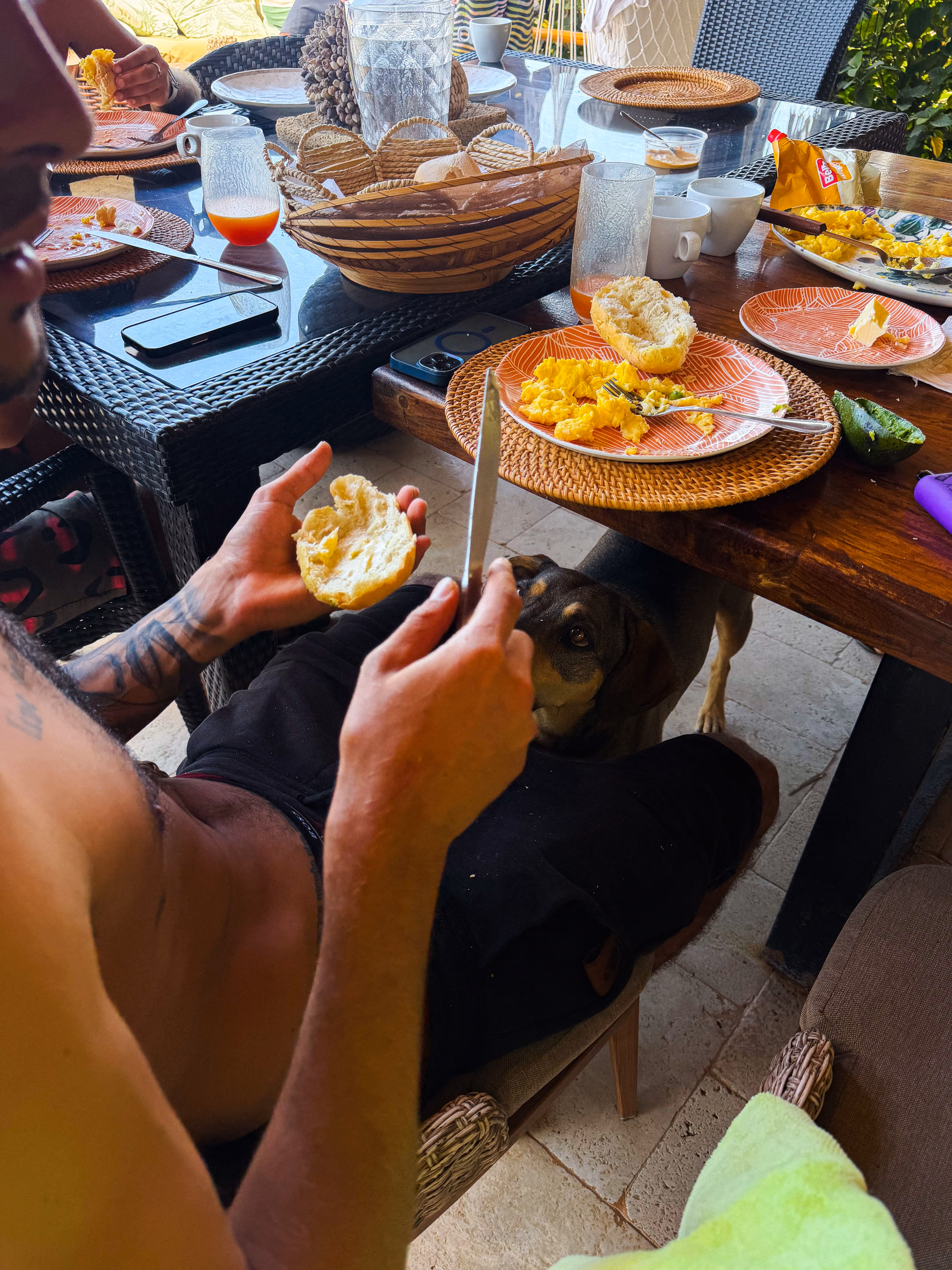 Man with tattoos buttering a bread roll at a breakfast table with scrambled eggs and a dog looking up.