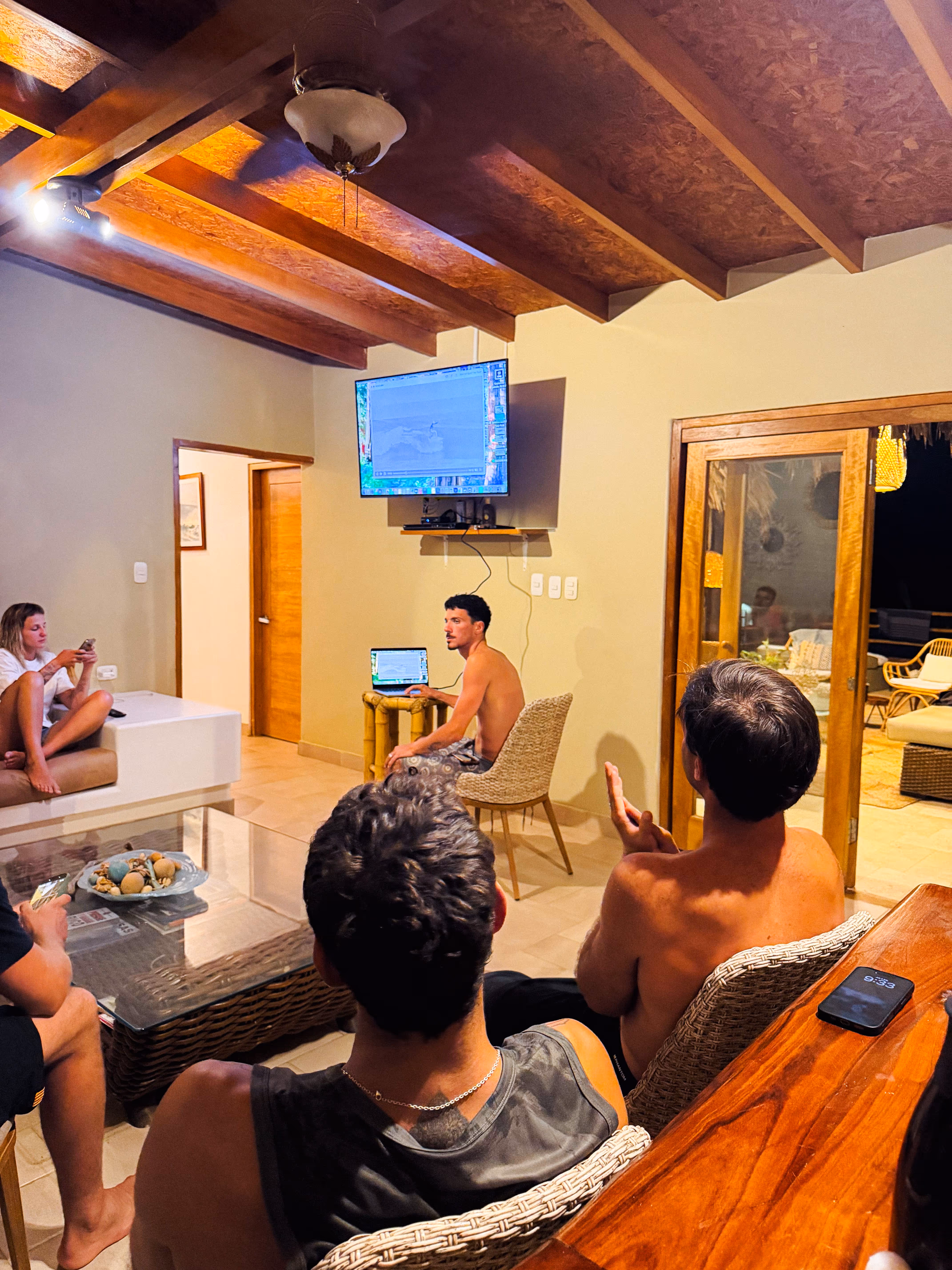 Group of people in a living room watching a shirtless man with a laptop presenting in front of a TV screen.