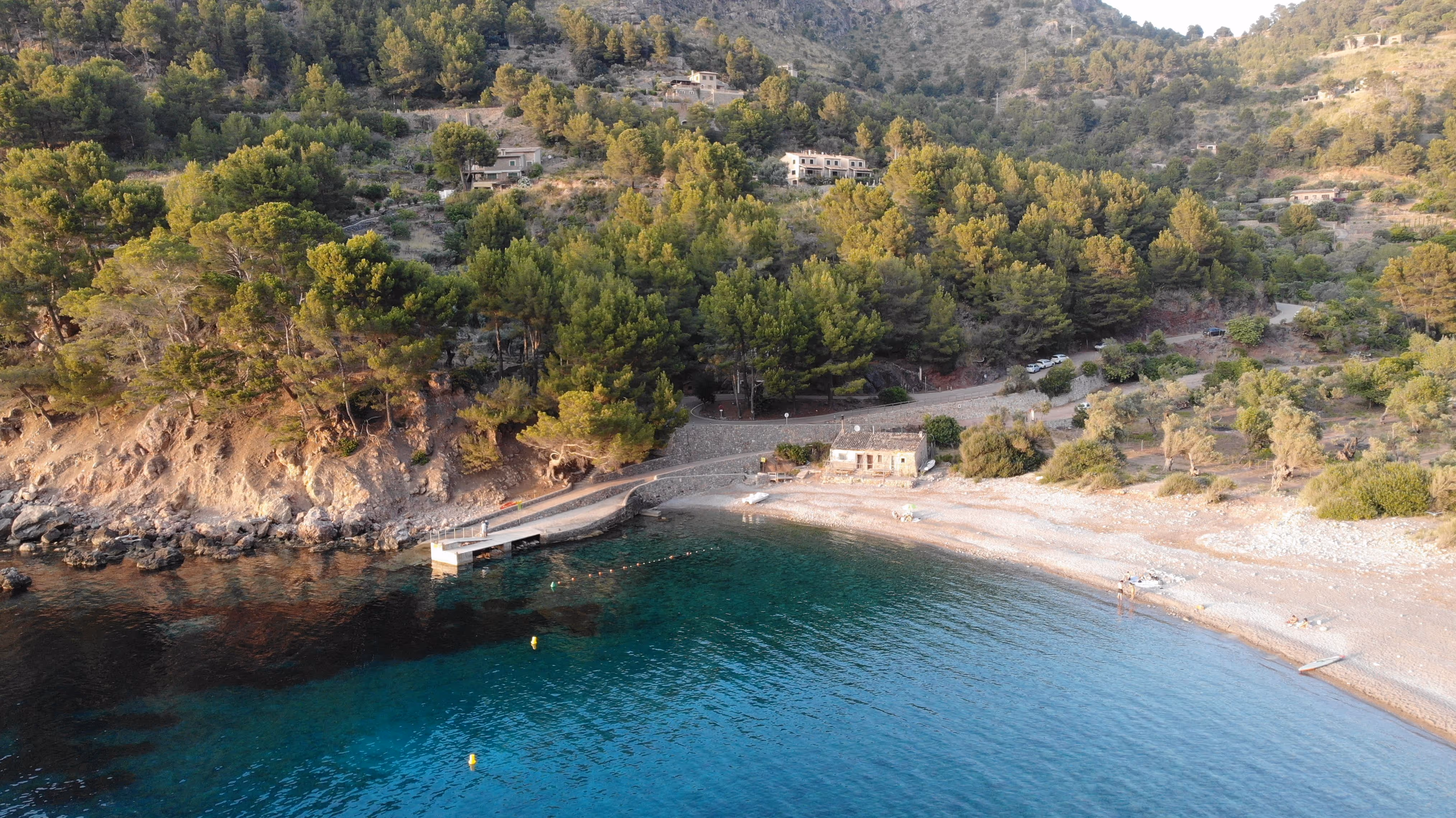 Aerial view of the pier at Cala Tuent with crystal-clear blue water and forested coastline.