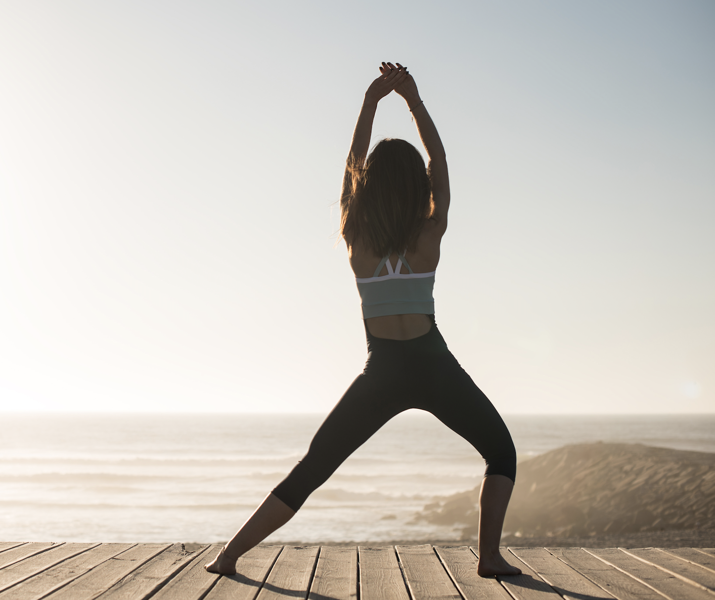 Woman in athletic wear practicing yoga on a wooden deck near the ocean at sunrise.