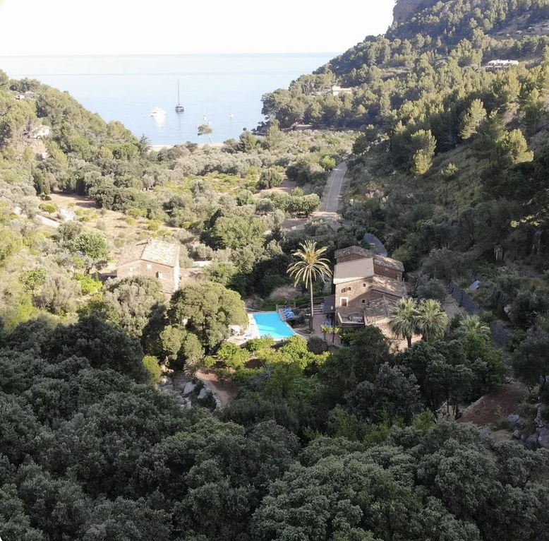 View from Finca Higo overlooking a lush green valley with terraced stone houses, a pool, and a distant view of the beach and sea at sunset.