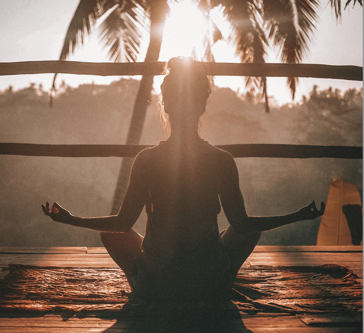 Silhouette of a person meditating in lotus position on a wooden deck with palm trees and sunrise in the background.