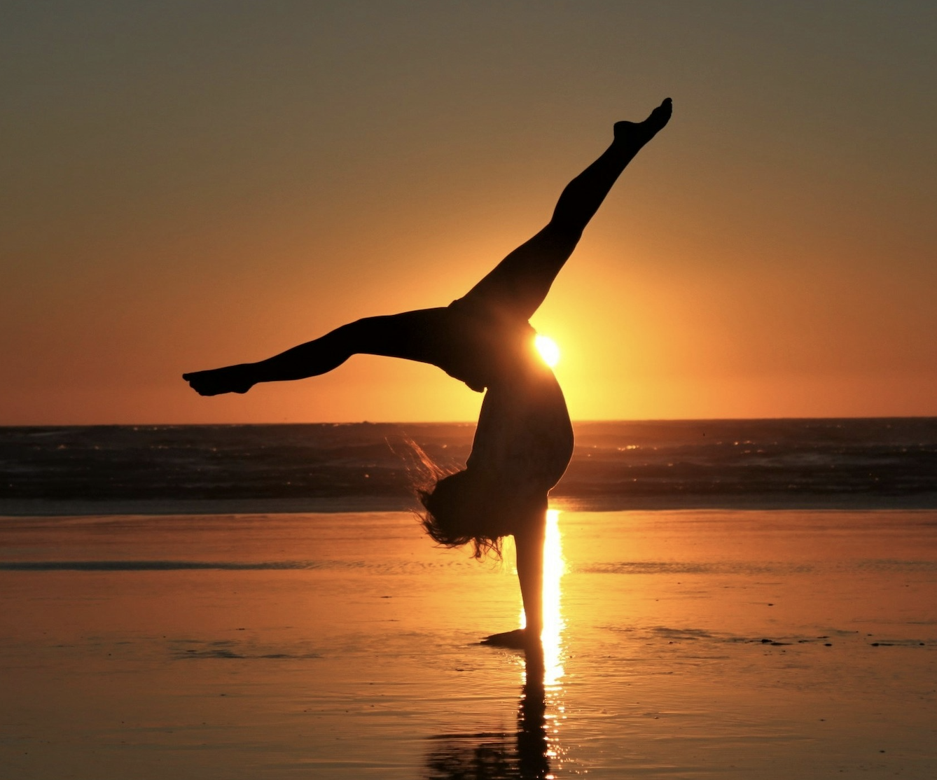 Silhouette of a person doing a handstand on a beach at sunset with the sun glowing behind them.
